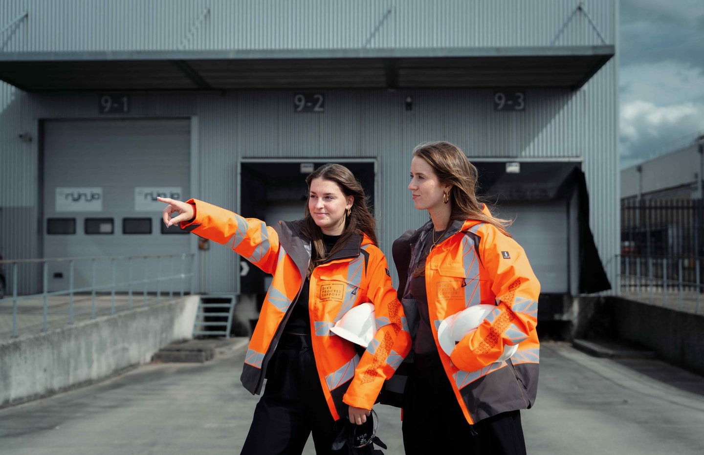 Twee vrouwen in oranje veiligheidsjas wijzen naar de laaddocks van een bedrijfshal