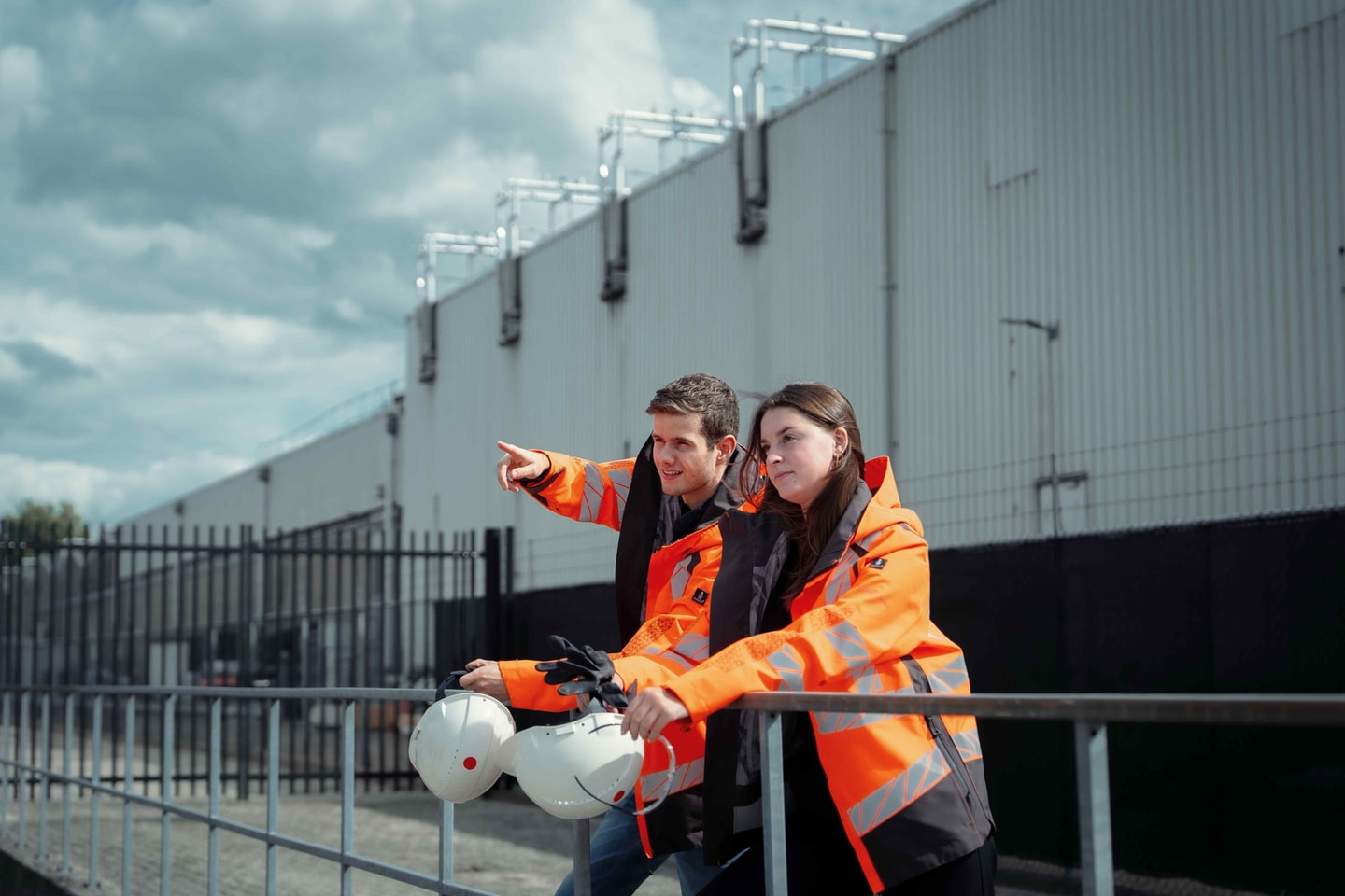 Man en vrouw in oranje jas met een helm in de hand wijzen langs een bedrijfshal
