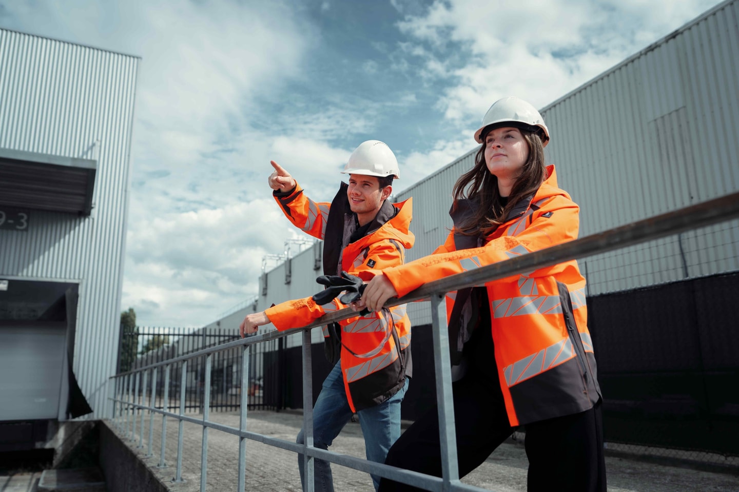Man en vrouw met helm en oranje jas wijzen vanaf een dakrand over een bedrijfsterrein