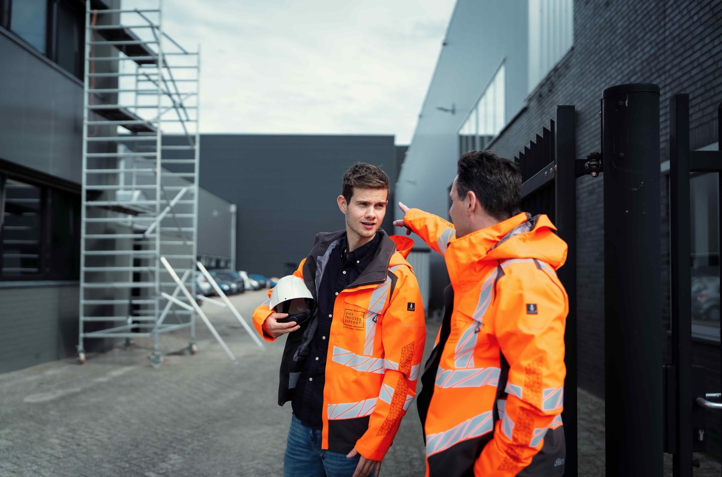 Twee mannen in oranje veiligheidsjas bij een steiger naast een bedrijfspand