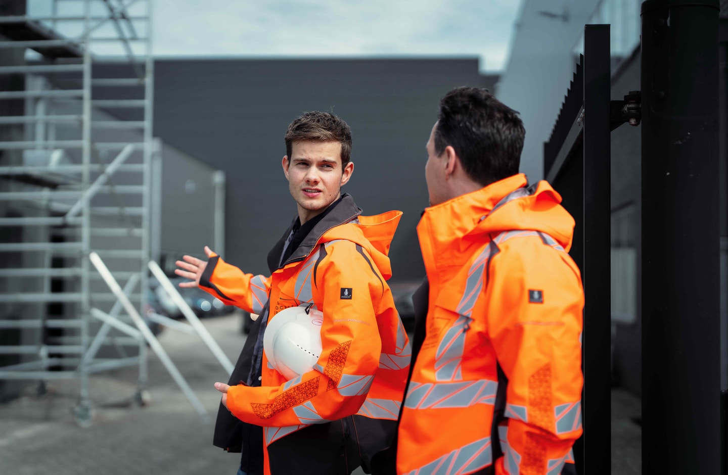Twee mannen in oranje jassen voor een steiger, een van hen met een witte helm in de hand