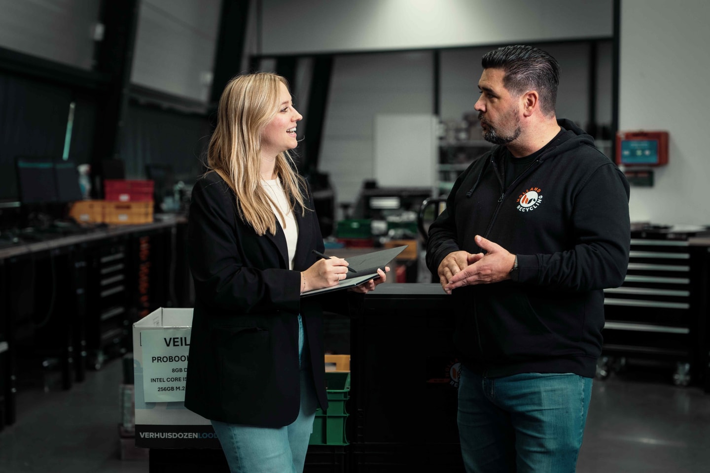 Vrouw met tablet in gesprek met een man in zwarte hoodie in een werkplaats