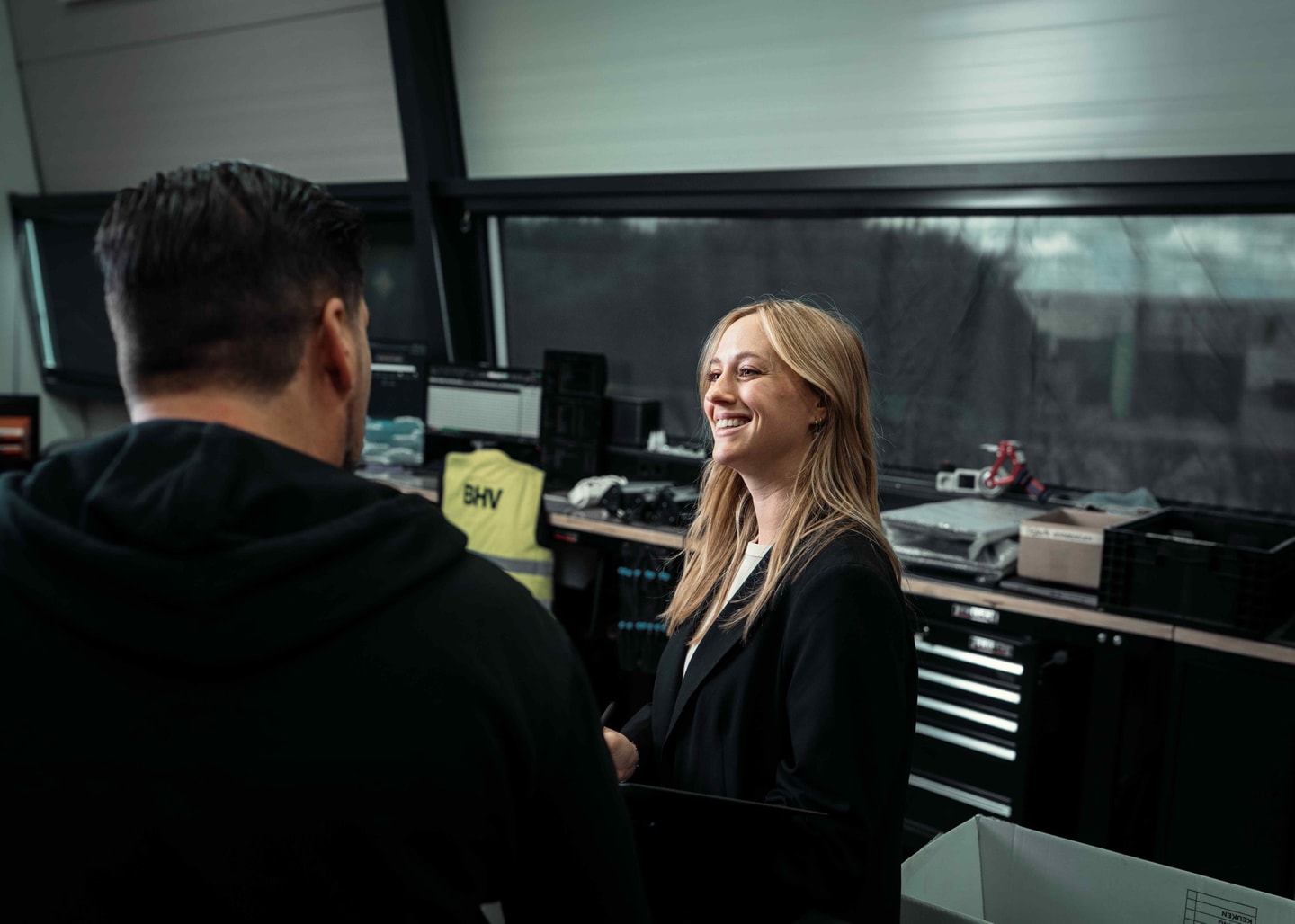 Vrouw lacht in gesprek met een man in een werkplaats met elektronica op de werkbanken
