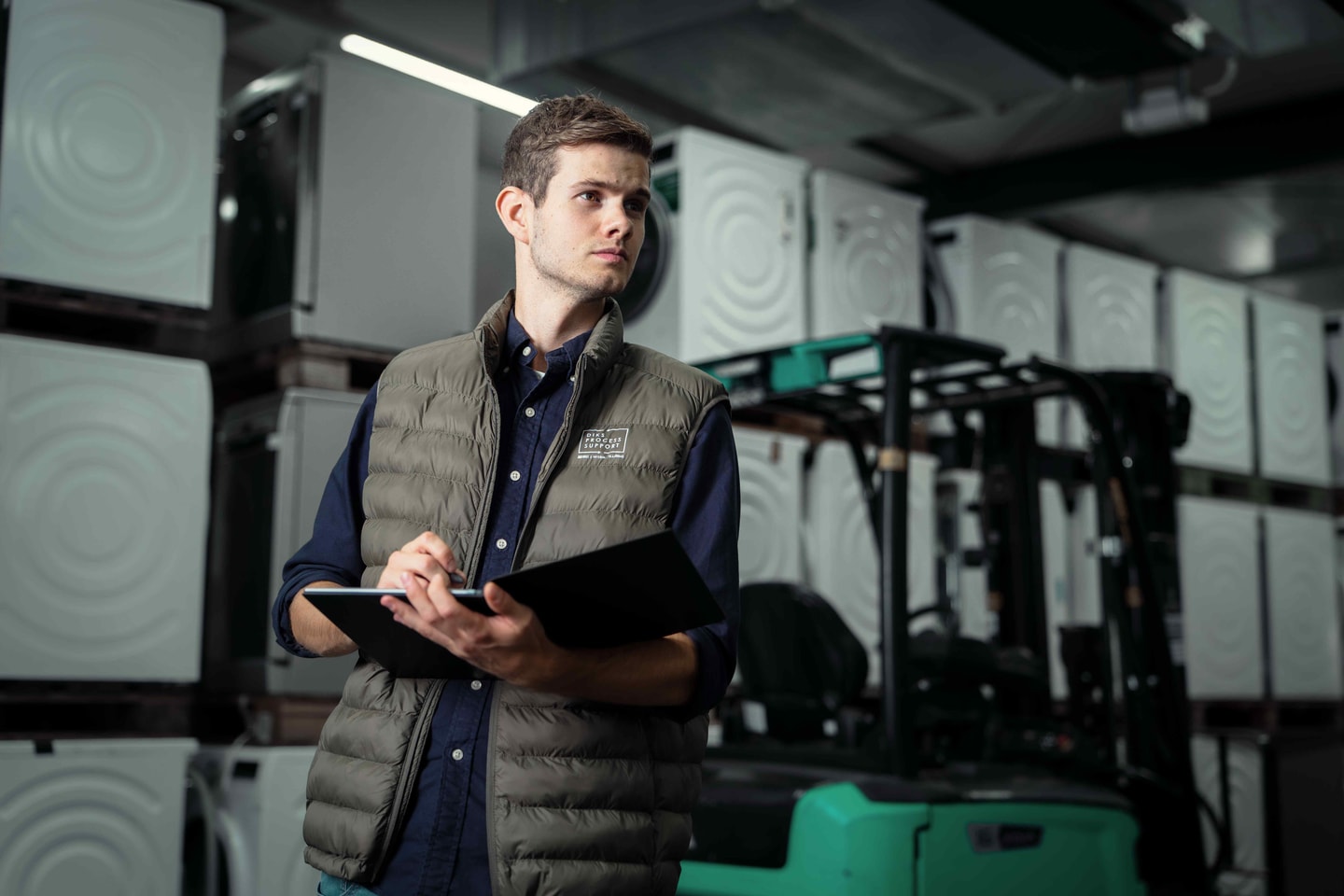 Man in bodywarmer met een map in een magazijn, met een groene heftruck achter hem