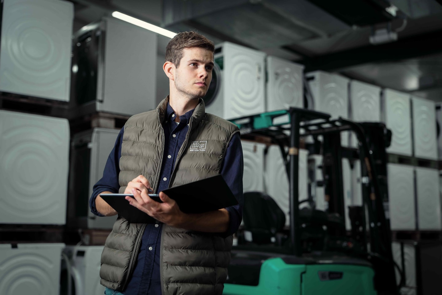 Man met bodywarmer en map tussen witte apparaten en een heftruck in een magazijn