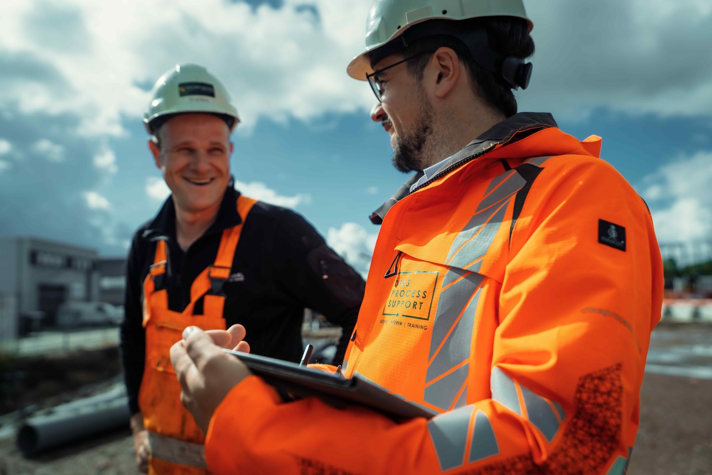 Twee mannen met helm en oranje werkkleding lachen in gesprek, één met een tablet