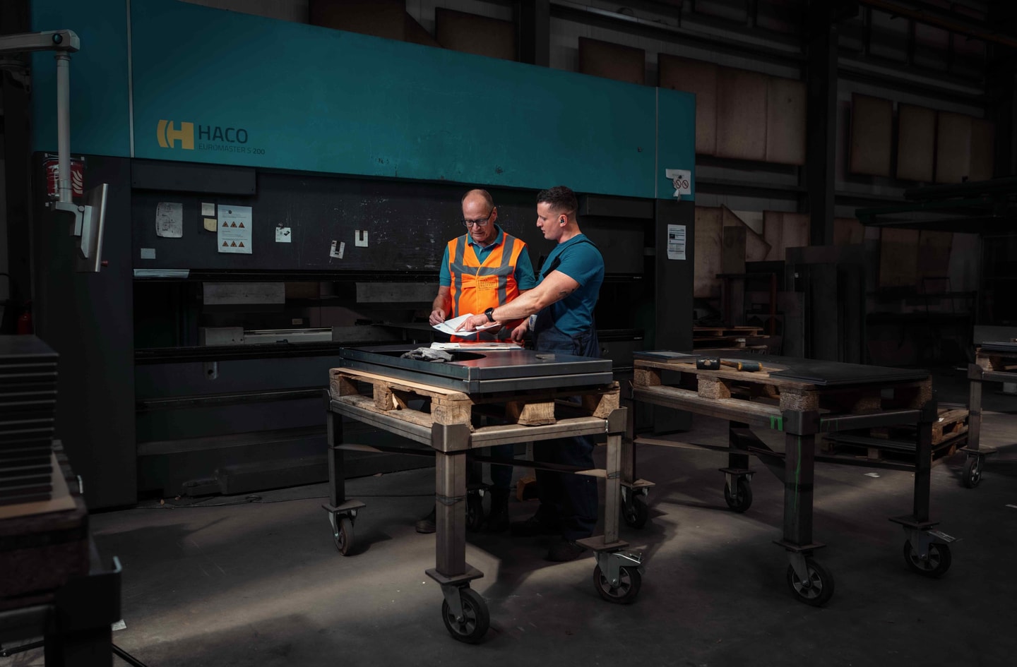 Twee mannen bekijken papieren aan een werktafel voor een kantbank in een metaalwerkplaats