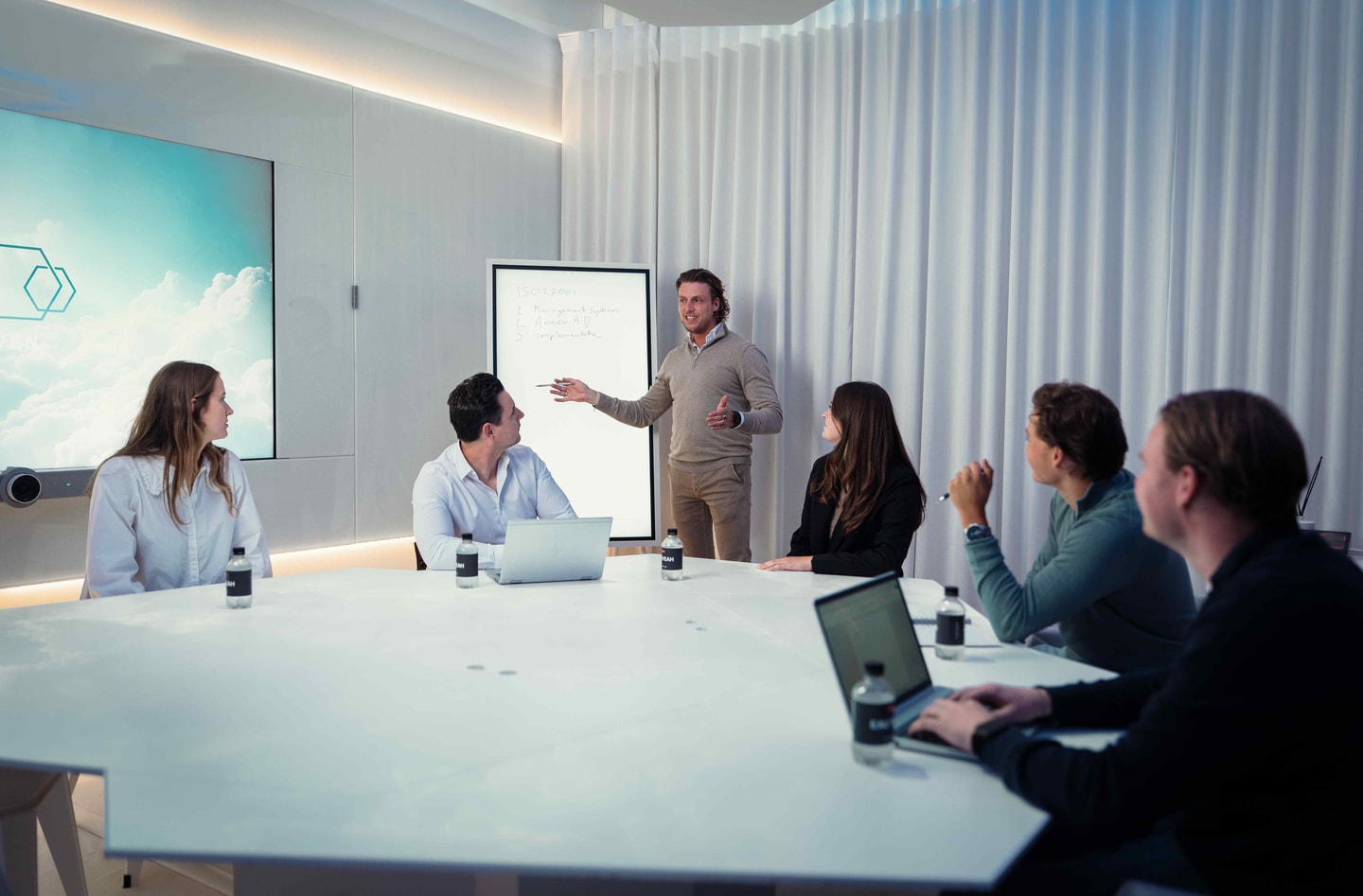 Man presenteert bij een whiteboard voor vijf mensen aan een witte vergadertafel