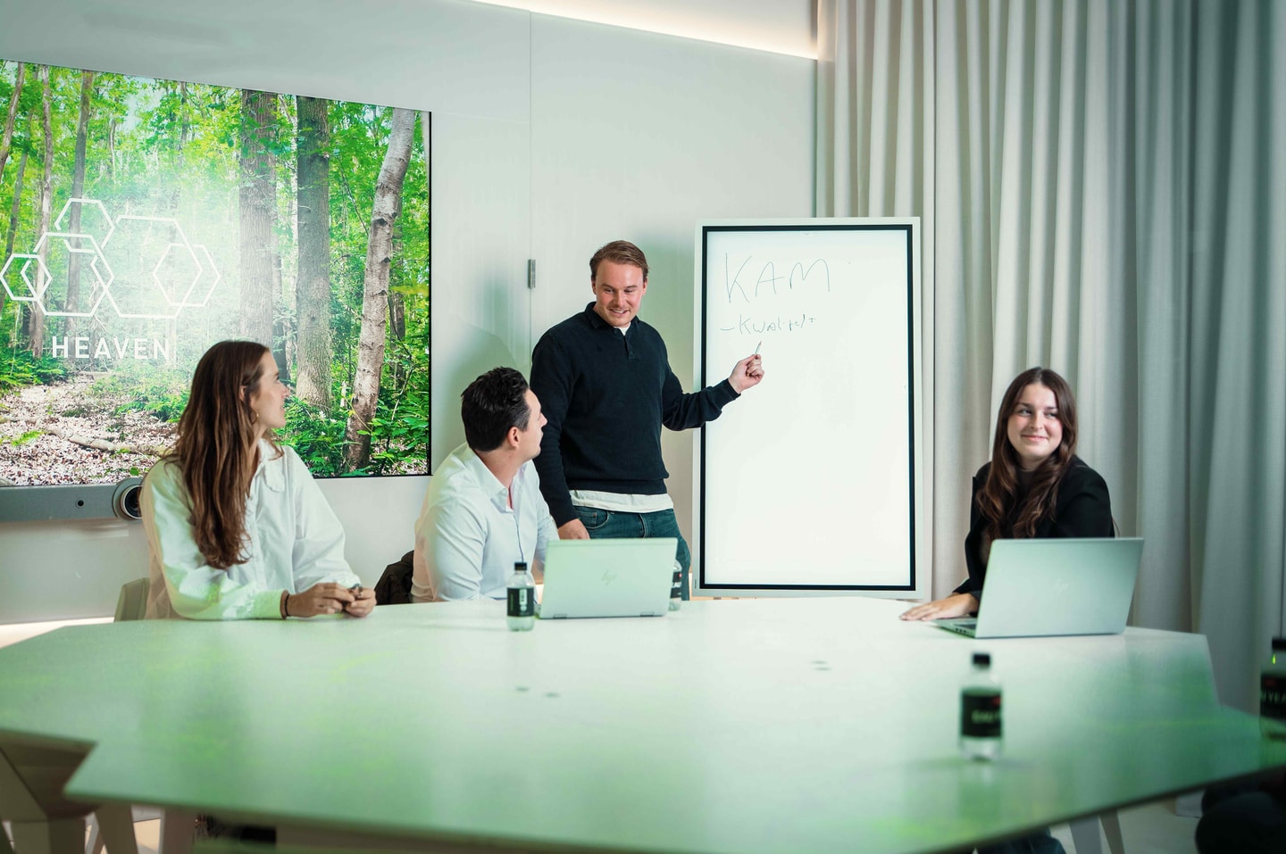 Een man geeft uitleg bij een whiteboard aan drie mensen die met laptops aan een vergadertafel zitten.