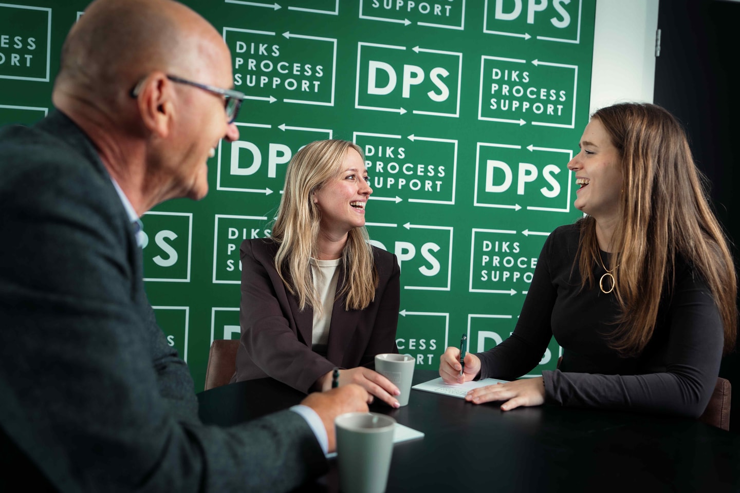 Drie mensen lachen aan een tafel voor de groene wand met het DPS-logo
