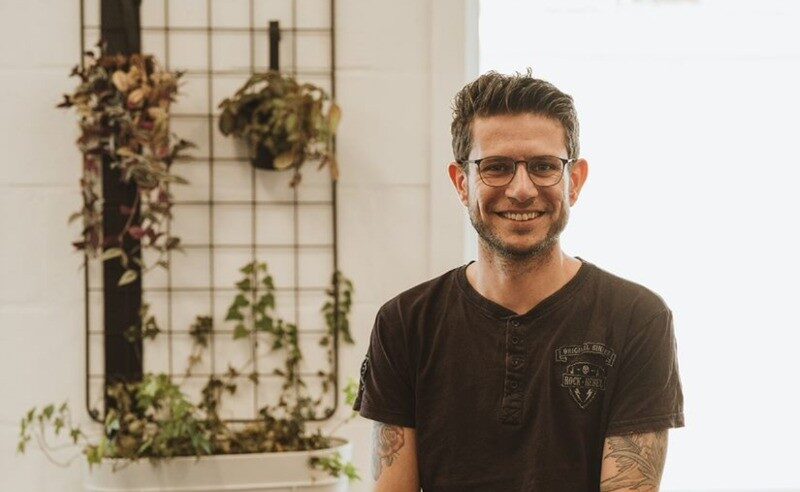 Lachende man met bril en tatoeages voor een wand met hangplanten