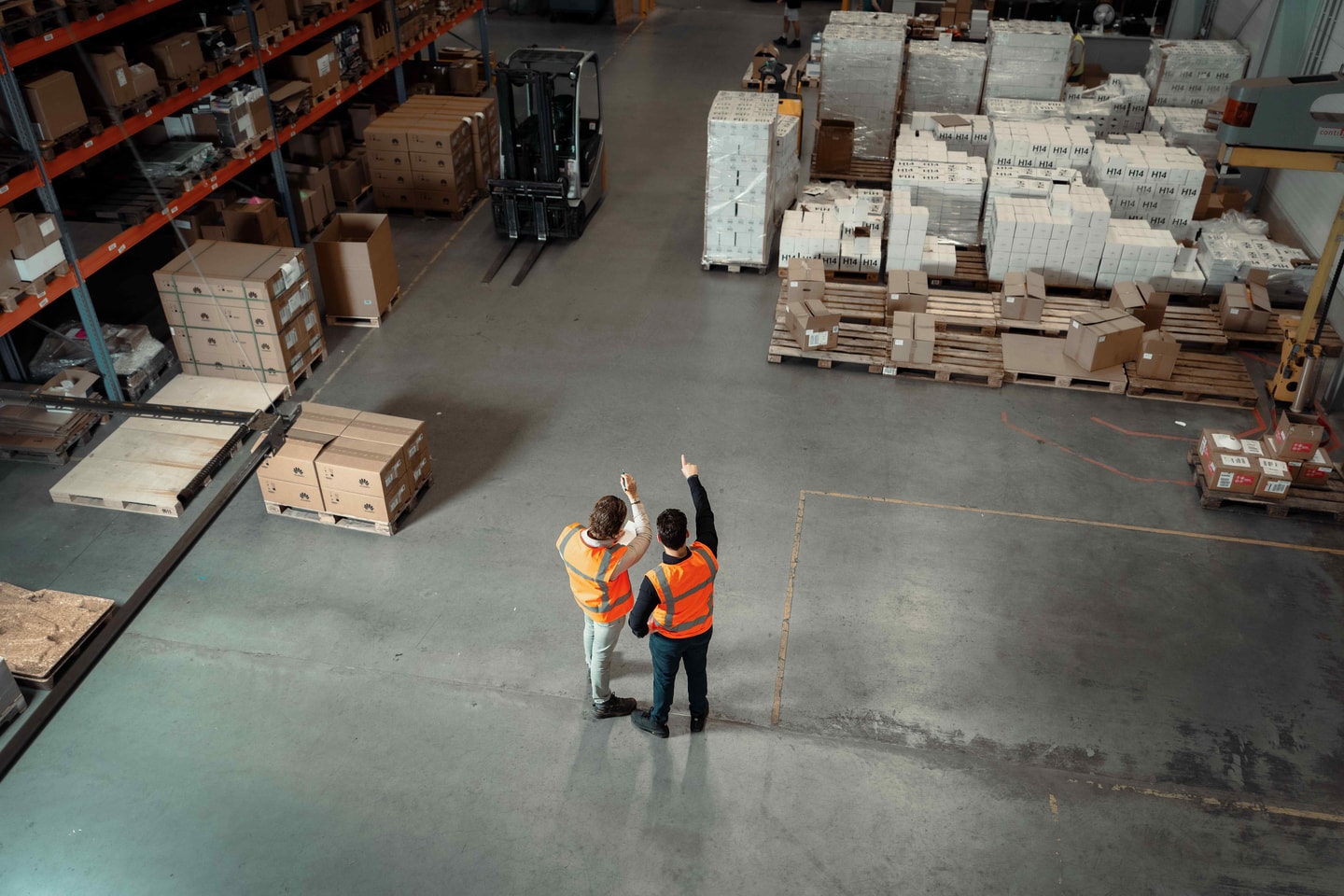 Twee medewerkers in oranje hesjes wijzen iets aan in een magazijn met pallets