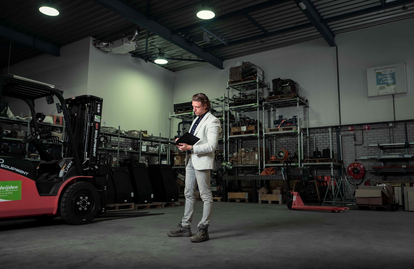 Man met een map tussen de stellingen van een werkplaats, met een heftruck ernaast